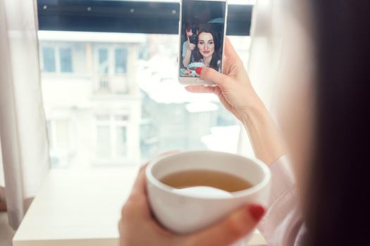 Woman taking a selfie picture in her bedroom holding a cup of tea