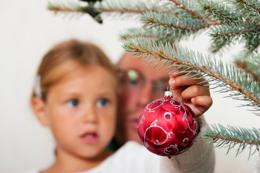 Young girl helping her father decorating the Christmas tree, holding some Christmas baubles in her hand (Focus on bauble)