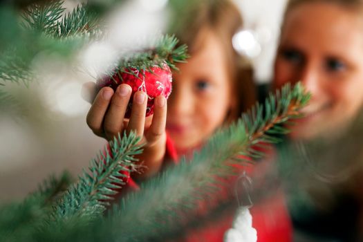 Young girl helping her mother decorating the Christmas tree, holding some Christmas baubles in her hand (Focus on bauble)