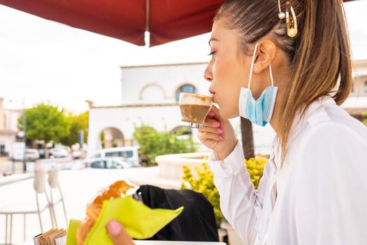 Beautiful woman profile portrait having breakfast in a bar terrace drinking cappuccino and eating brioche with lowered protective mask. New normal city life at Coronvirus times: business woman break