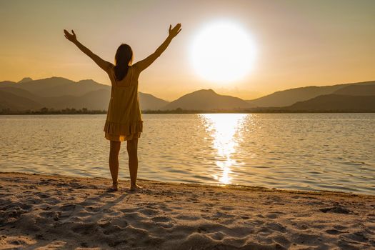 Suggestive shot of rear view of thoughtful woman silhouette in spiritual pose with open arms looking at sunset or sunrise barefoot on sea sand or ocean beach. Find yourself in contact with nature