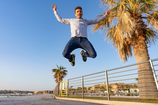 Happy boy jumping high with open arms looking at camera thinking about success in life. The energy of positive thinking for a better life. Young man smiling celebrate goal achieved with work or study