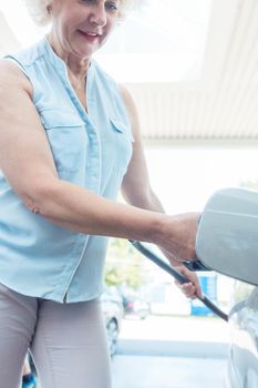 Low-angle view portrait of an active senior woman smiling while filling up the gas tank of her car at the station in summer