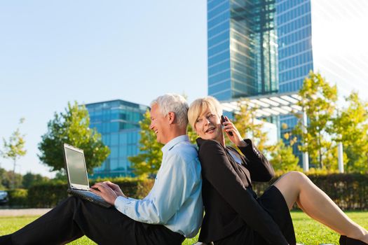 Business people working outdoors on a meadow - he is working with laptop, she is calling someone on phone