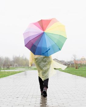 woman in yellow raincoat holding rainbow umbrella out in the rain. People from behind, rear view
