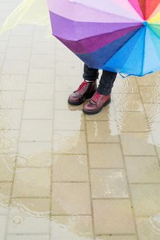 woman in red shoes holding rainbow umbrella out in the rain, standing in a puddle