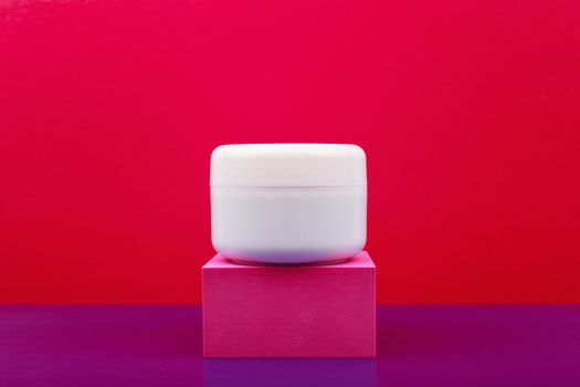 White glossy cosmetic jar on pink pedestal against red and purple background with copy space. Concept of beauty product, cream, mask, balm or scrub