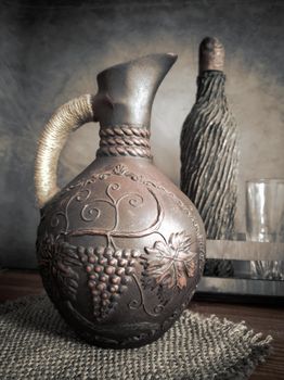 Ceramic wine jug made of red clay with a braided handle, handmade. There is a wine bottle and a glass next to it.