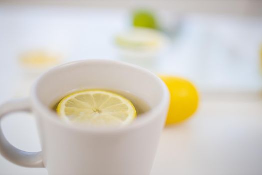 Close-up of cup of lemon tea on white table with blurry limes and lemons as background. White mug with lemon slice floating inside. Sweet citric beverages