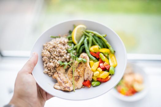 Hand holding chicken and buckwheat dish with green beans, broad beans, tomato, and pepper slices. Nutritious dish with vegetables and meat on white plate. Healthy balanced diet