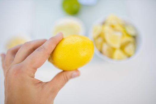 Top view of female hand holding a lemon above blurry small bowl of lime and lemon slices. Woman cutting lemons into small pieces. Citrus drink preparation