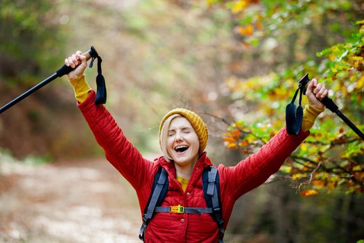 Hiking girl with poles and backpack on a trail. Hands up enjoying in nature. Travel and healthy lifestyle outdoors in fall season.