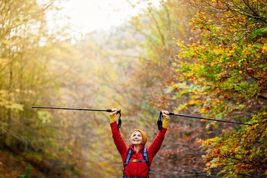 Hiking girl with poles and backpack on a trail. Hands up enjoying in nature. Travel and healthy lifestyle outdoors in fall season.