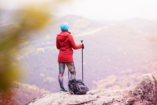 Hiking girl with poles and backpack standing on rocks. Windy autumn day. Travel and healthy lifestyle outdoors in fall season