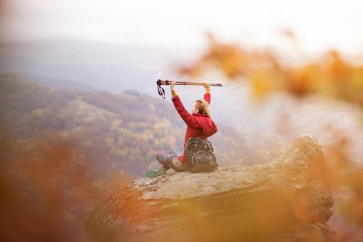 Hiker girl sitting on a rock in the mountains. Enjoying the view with hands up holding hiking poles. Travel and healthy lifestyle outdoors in fall season