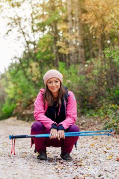 Hiker girl crouching on a wide trail in the mountains. Backpacker with pink jacket in a forest. Healthy fitness lifestyle outdoors.