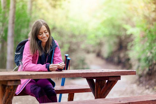 Hiker girl resting on a bench in the forest. Backpacker with pink jacket holding dry autumn leaves.
