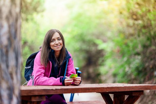 Hiker girl resting on a bench in the forest. Backpacker with pink jacket holding dry autumn leaves.