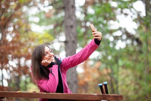 Hiker girl resting on a bench in the forest. Backpacker with pink jacket taking selfie with smartphone.