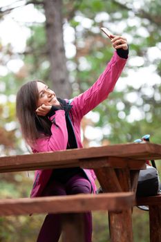 Hiker girl resting on a bench in the forest. Backpacker with pink jacket taking selfie with smartphone.