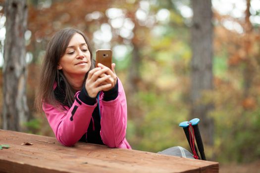 Hiker girl resting on a bench in the forest. Backpacker with pink jacket holding cell phone.