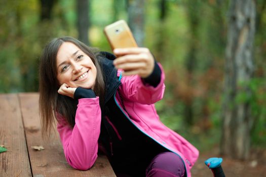 Hiker girl resting on a bench in the forest. Backpacker with pink jacket taking selfie with smartphone.