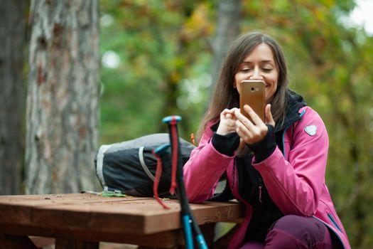 Hiker girl resting on a bench in the forest. Backpacker with pink jacket holding cell phone.