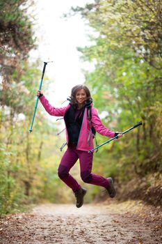 Hiker girl jumping on a trail in the mountains. Backpacker with hiking poles and pink jacket in a forest. Happy lifestyle outdoors.