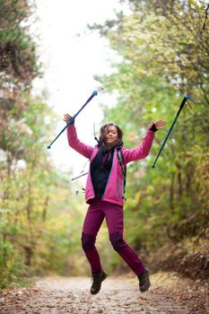 Hiker girl jumping on a trail in the mountains. Backpacker with hiking poles and pink jacket in a forest. Happy lifestyle outdoors.
