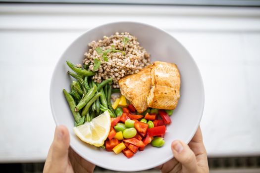 Top view of hands holding salmon and buckwheat dish with green beans, broad beans, and tomato slices. Nutritious dish with vegetables and fish on white plate. Healthy balanced diet