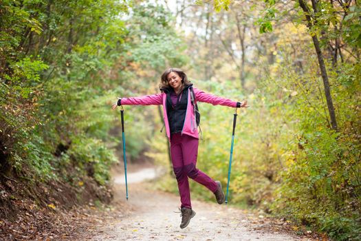 Hiker girl jumping on a trail in the mountains. Backpacker with hiking poles and pink jacket in a forest. Happy lifestyle outdoors.