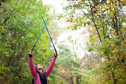 Hiking girl with poles and backpack on a trail. Hands up enjoying in nature. Travel and healthy lifestyle outdoors in fall season.