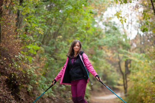 Hiker girl jumping on a trail in the mountains. Backpacker with hiking poles and pink jacket in a forest. Happy lifestyle outdoors.