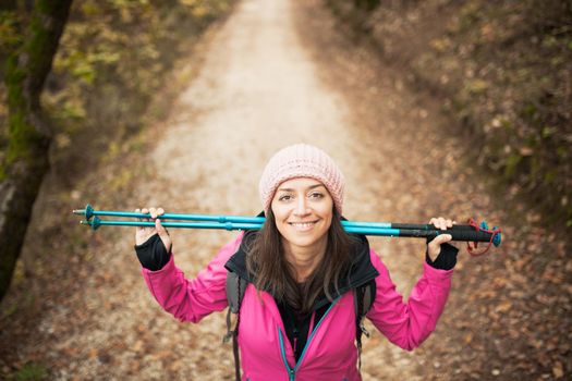 Hiker girl in pink on a trail in the forest. Looking at camera with poles in hands. Nature in fall season.