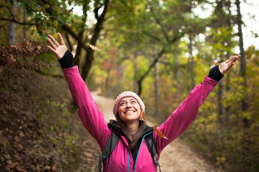 Hiking girl in pink on a trail in the forest. Hands up enjoying the falling leaves in nature in fall season.