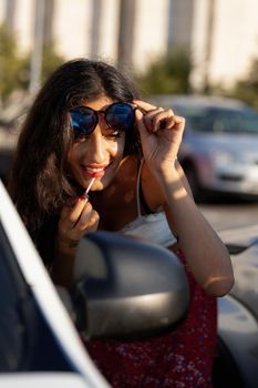 Beautiful girl with sunglasses putting on lipstick using a car mirror. Sunny summer day.