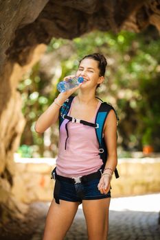 young and happy hiker girl drinking water from a plastic bottle next to a rocky passage