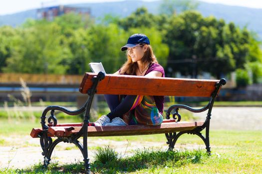 brunette girl with a cap, reading a book sitting on a bench