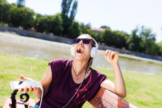 brunette girl with headphones and smartphone smiling while sitting on a bench