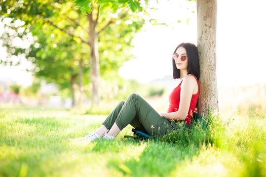 girl with dark brown hair and red sunglasses posing under a tree