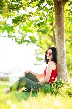 girl with dark brown hair and red sunglasses posing under a tree