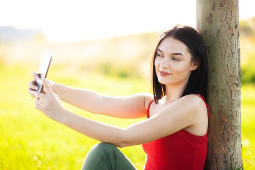 girl with dark brown hair using smartphone taking selfy under a tree