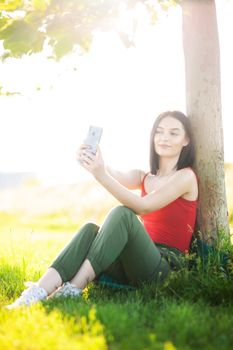girl with dark brown hair using smartphone taking selfy under a tree