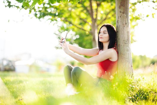 girl with dark brown hair using smartphone taking selfy under a tree