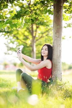 girl with dark brown hair using smartphone taking selfy under a tree
