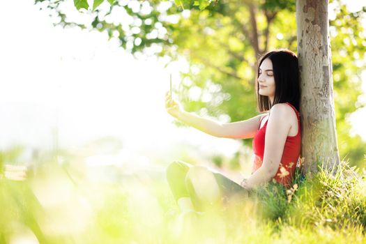 girl with dark brown hair using smartphone taking selfy under a tree