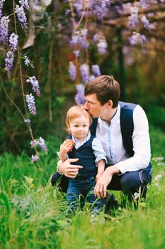 Father is kissing his baby boy on top of the head with a wysteria blooming in the background. High quality photo