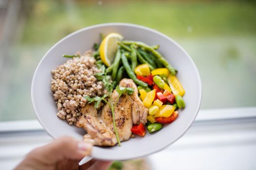 Hand holding chicken and buckwheat dish with green beans, broad beans, tomato, and pepper slices. Nutritious dish with vegetables and meat on white plate. Healthy balanced diet