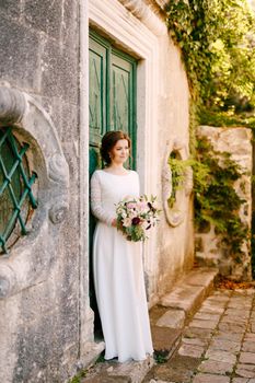 Smiling bride stands against the background of a building with a beautiful bouquet of flowers in her hands. High quality photo