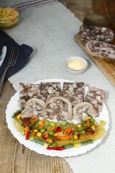 On the table on a white plate is sliced pork stomach, stuffed with meat and bacon, next to a variety of vegetables. It's a delicious meat snack.